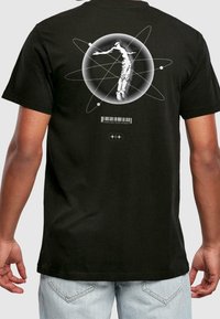 Zwart katoenen T-shirt met een grafisch ontwerp van een figuur binnen een cirkelvormig atoommotief, met daaronder een streepjescode en de tekst "MAN ON THE MOON".