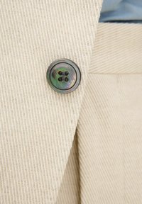 Beige kordfärg med vertikala ribbor. Har en rund, mörk knapp med en iriserande finish och fyra hål för fäste.