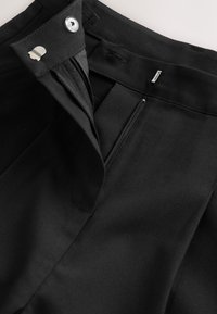 Vita di pantaloni neri con cerniera aperta, bottone e dettaglio di chiusura con gancio in metallo.