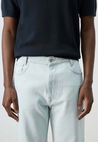 Persona que lleva una camiseta negra texturizada de manga corta metida en unos vaqueros vaqueros de color azul claro, de pie con los brazos relajados a los lados.