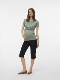 Grūtniece, kas ģērbusies zaļā ribotā īsās piedurkņu topā, melnos ceļgalu garumā biksēs un brūnās naturālajās kurpēs, stāvot pret vienkāršu fona.