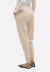 Mujer vestida con pantalones de chándal beige ajustados, con bolsillos laterales y zapatos de punta negra con tiras en los tobillos, de pie de lado sobre un fondo blanco.