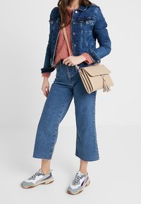 Giacca di jeans con chiusura a bottoni, blu chiaro; maglione di maglia rosa; jeans a gamba larga; borsa a tracolla beige con frange; sneakers grigie e bianche.