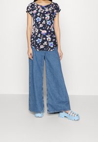 Haut floral marine à manches courtes, associé à un jean bleu clair à jambes larges et à des sandales bleu chunky avec un design ouvert et une semelle épaisse.