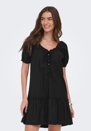 Ung kvinde med langt brunt hår iført en kort sort kjole med pufærmer og knapdetaljer, smilende og kiggende til siden.