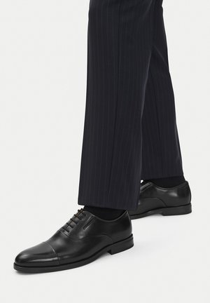 Czarne skórzane buty wizytowe o zaokrąglonym nosku, z tonowaną nicią i gładkim wykończeniem, noszone z spodniami w prążki.