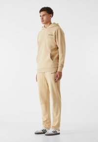 Felpa beige con cappuccio e jogger abbinati realizzati in un tessuto morbido, caratterizzati da una tasca a marsupio e un logo in rilievo sul petto e sulla coscia.