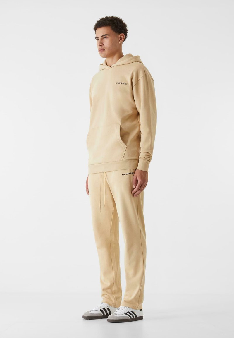 Beige hoodie och matchande joggers i mjukt tyg, med kenguruficka och präglad logotyp på bröstet och låret.