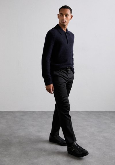 Junger Mann, der drinnen geht, trägt einen schwarzen Langarmpullover, schwarze Hosen mit Gürtel und schwarze Sneakers vor einer schlichten hellgrauen Wand.