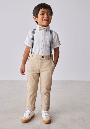 Jonge jongen glimlacht, draagt wit overhemd, beige strikje, bretels, beige broek en witte sneakers, staand op een lichtgrijze vloer.