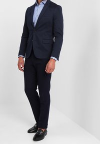 Granatowy, dopasowany garnitur z dopasowanymi ciemnymi spodniami, jasnoniebieską koszulą w wzory oraz czarnymi butami wsuwanymi. Prosty i uporządkowany fason.