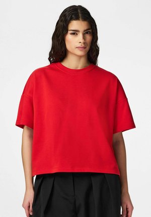 Kvinde med langt, mørkt, bølget hår iført en løs, rød top med korte ærmer og sorte plisserede bukser, stående mod en ensfarvet hvid baggrund.