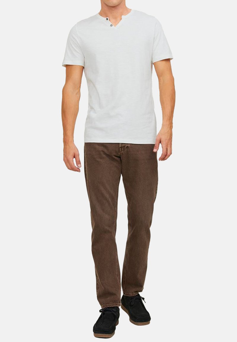 Weißes Kurzarm-Henley-T-Shirt mit V-Ausschnitt und zwei Knöpfen, kombiniert mit braunen Jeans und schwarzen Schnürschuhen.