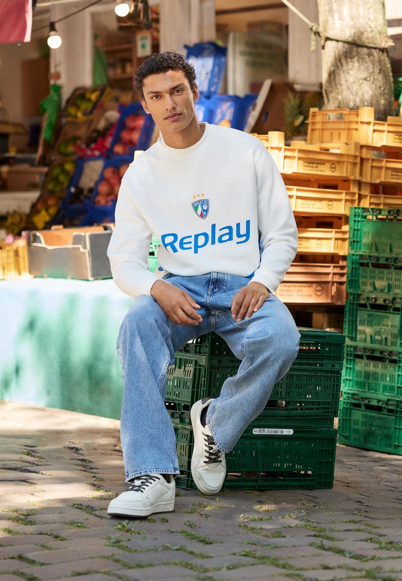 Hvit hettegenser med blå "Replay" logo og blå-grønn emblem. Lyseblå jeans, hvite sneakers med svarte detaljer. Sitter på grønne kasser.