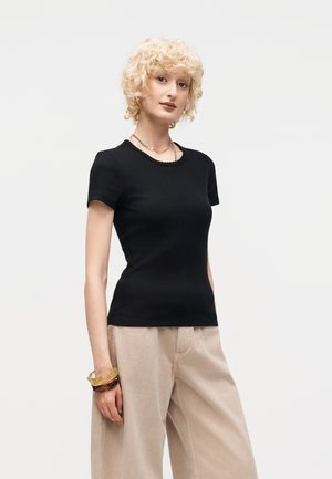 Νεαρό άτομο με σγουρά ξανθά μαλλιά που φοράει εφαρμοστό μαύρο τοπ με κοντά μανίκια, μπεζ παντελόνι φαρδιάς γραμμής και χοντρά βραχιόλια στον αριστερό καρπό.