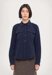 Navy blue button-up shirt with a collar, two front pockets, and shoulder epaulettes. Soft, smooth fabric with a relaxed fit, long sleeves.
