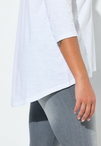 Haut blanc ample à manches trois-quarts, présenté en angle. Associé à un jean gris clair ajusté, avec une texture subtile.