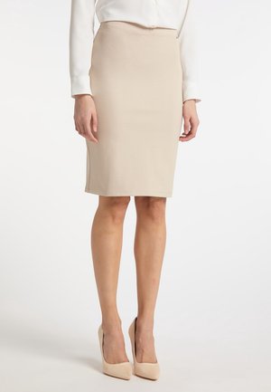 Femme portant une jupe crayon beige jusqu'aux genoux associée à des escarpins beiges et un chemisier blanc à manches longues, debout sur un fond neutre.