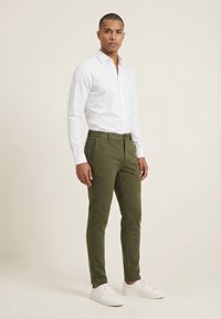 Homme debout portant une chemise blanche boutonnée, un pantalon slim vert olive et des baskets blanches sur un fond clair uni.