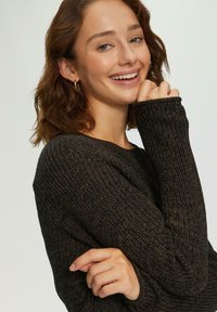 Gebreide trui in donkerbruin, met een gestructureerd patroon, ronde hals en lange mouwen met geribbelde manchetten. Model lacht, hand dicht bij het gezicht.