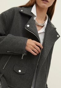 Veste en mélange de laine grise avec col à cran, fermeture éclair argentée et boutons-pression. Comprend une poche poitrine et une poche latérale zippée.
