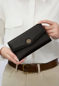 Portafoglio in pelle nera con texture, chiusura a pattina e hardware dorato con logo, tenuto in mano contro una camicia bianca e pantaloni beige.