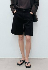 Shorts en denim noir à coupe droite, munis de poches latérales et de passants de ceinture. Portés avec un haut sombre et des tongs noires. Design minimaliste.