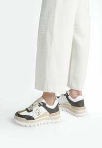 Las zapatillas presentan una combinación de materiales en marrón oscuro, blanco y dorado, con un diseño estampado, suela gruesa y acentos decorativos de logo.