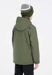 Giacca impermeabile verde con cappuccio, polsini elastici e un logo sulla spalla. Abbinata a un berretto a maglia a righe in grigio e verde.