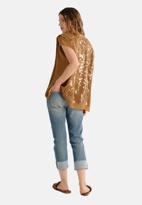 Top in bronzo senza maniche con dettagli in paillettes sulla schiena, abbinato a jeans azzurri con risvolto e sandali marroni. Tessuto morbido e vestibilità comoda.