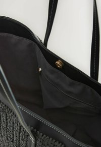 Sac fourre-tout noir avec texture tissée et accents en cuir lisse, s'ouvrant pour révéler un intérieur spacieux avec une poche zippée.