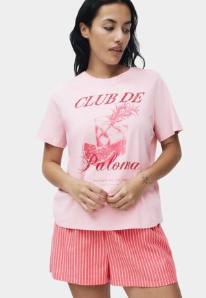 Vrouw met een roze T-shirt met een rood cocktail-illustratie en tekst, gecombineerd met rood-wit gestreepte korte broek, staand tegen een lichte achtergrond.