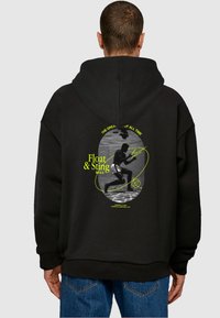 Hanorac oversized negru, cu un imprimeu grafic al unui înotător, evidențiat de accente neon galbene și text. Material moale, mâneci lungi și glugă.