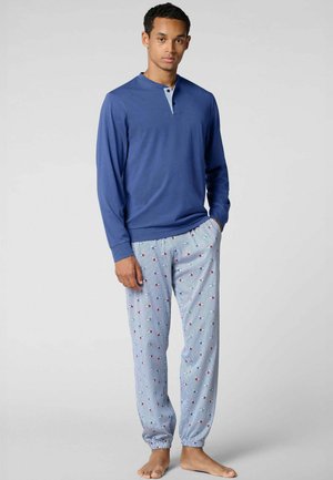 Giovane uomo che indossa una maglia henley a maniche lunghe blu e pantaloni del pigiama a righe azzurre chiare con piccoli motivi di barche rosse e bianche.