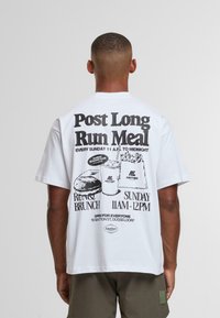 Hombre que lleva una camiseta blanca con estampado negro que promociona un brunch dominical de 11 AM a medianoche, con ilustración de hamburguesa, bebida y papas fritas.
