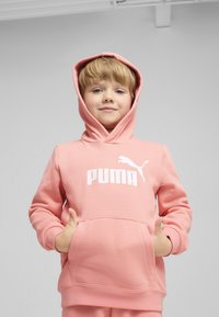 Rozā kapuce ar priekšējo kabatu, kurai ir balts "PUMA" logo. Mīksta tekstūra un brīva piegriezuma, piemērota ikdienas valkāšanai.