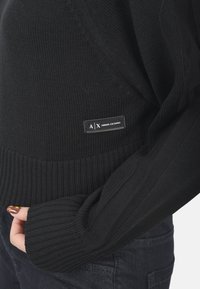 Maglione nero a maglia con polsini e orlo a coste. Presenta un piccolo etichetta con "A|X ARMANI EXCHANGE". Texture morbida, vestibilità casual e design minimal.