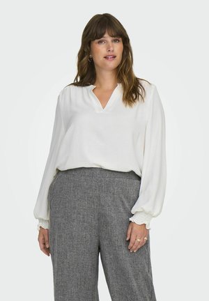 Haut blanc à manches longues avec décolleté en V et poignets froncés, associé à un pantalon large en tissu texturé gris. Fini en tissu lisse.