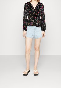 Blouse portefeuille noire à motifs floraux avec manches longues, associée à un short en denim bleu clair et des tongs noires à plateforme. Tissu lisse, motifs colorés.