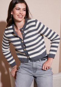 Ung kvinde smiler, iført en grå og hvid stribet cardigan med knapper og lysegrå højttaljede jeans, poserer indendørs foran en beige væg.