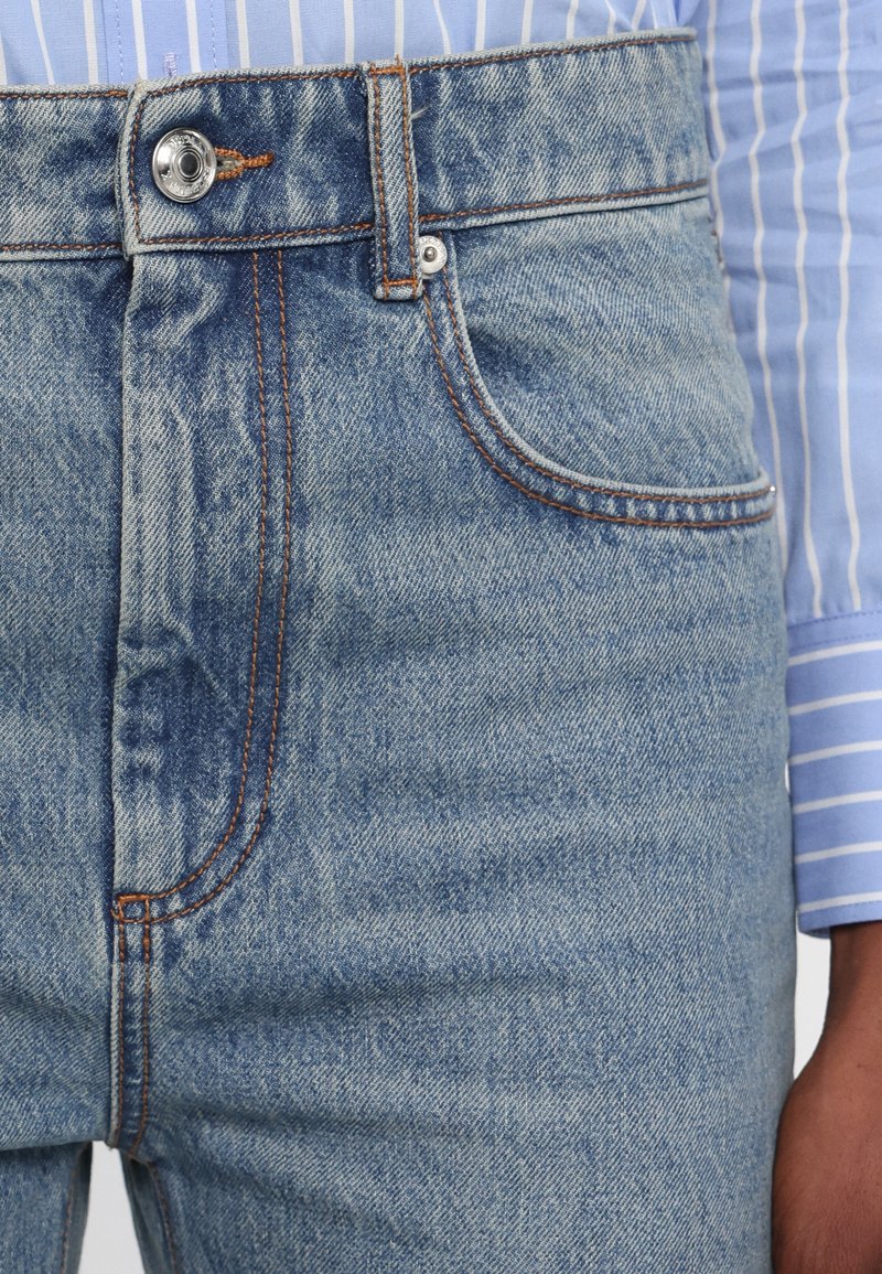 Light blue denim jeans with a visible button and zip closure, featuring orange stitching and a front pocket on the left side.