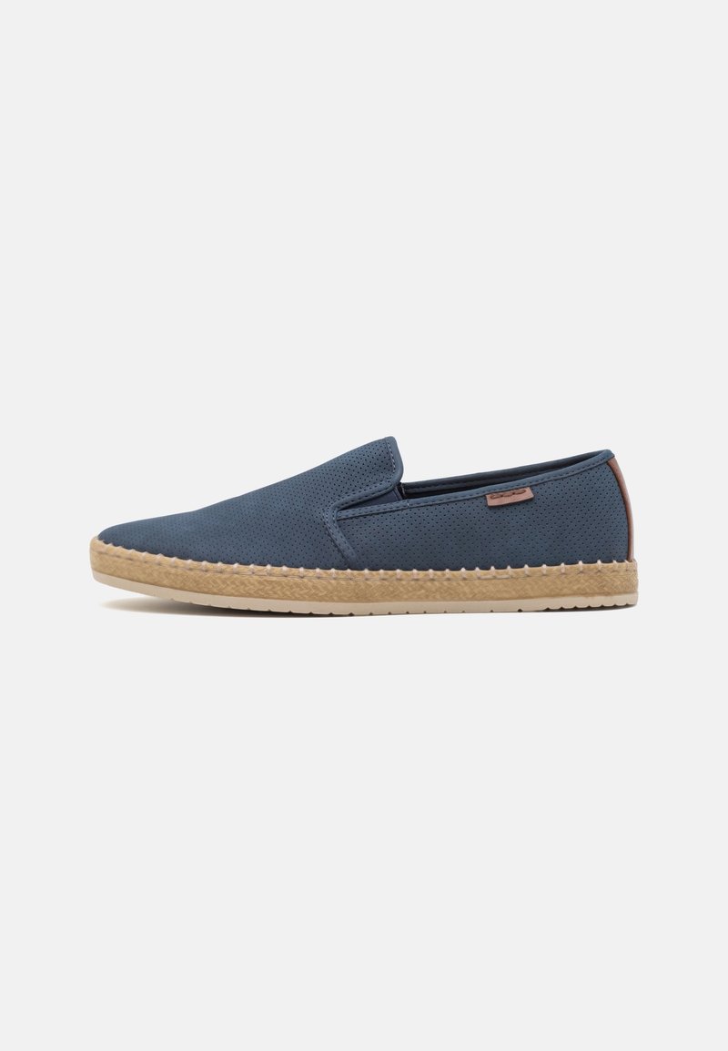 Une chaussure à enfiler bleu marine unique avec dessus perforé, semelle tissée beige et petite languette marron au talon sur fond blanc uni.