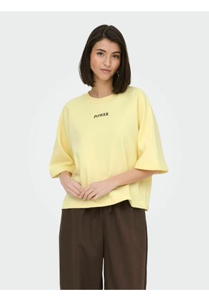 Moteris su tiesiais tamsiais plaukais, dėvinti laisvą geltoną džemperį su užrašu „POWER“ ir plačias rudas kelnes, stovinti priešais vienspalvį foną.