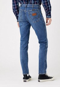 Jeans de ganga azul com ajuste slim, bolsos traseiros, detalhe em patch de couro e costura contrastante, combinados com ténis altos pretos.