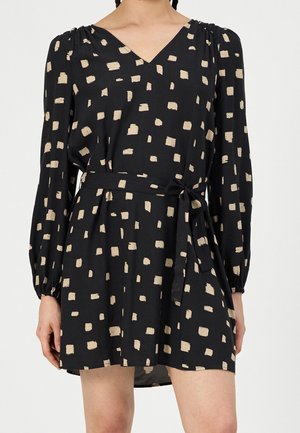 Femme portant une robe noire à manches longues avec des motifs carrés beige et une ceinture nouée à la taille, coupée au-dessus des genoux.