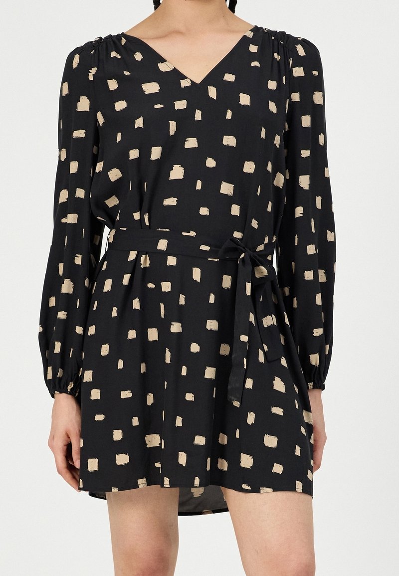 Femme portant une robe noire à manches longues avec des motifs carrés beige et une ceinture nouée à la taille, coupée au-dessus des genoux.