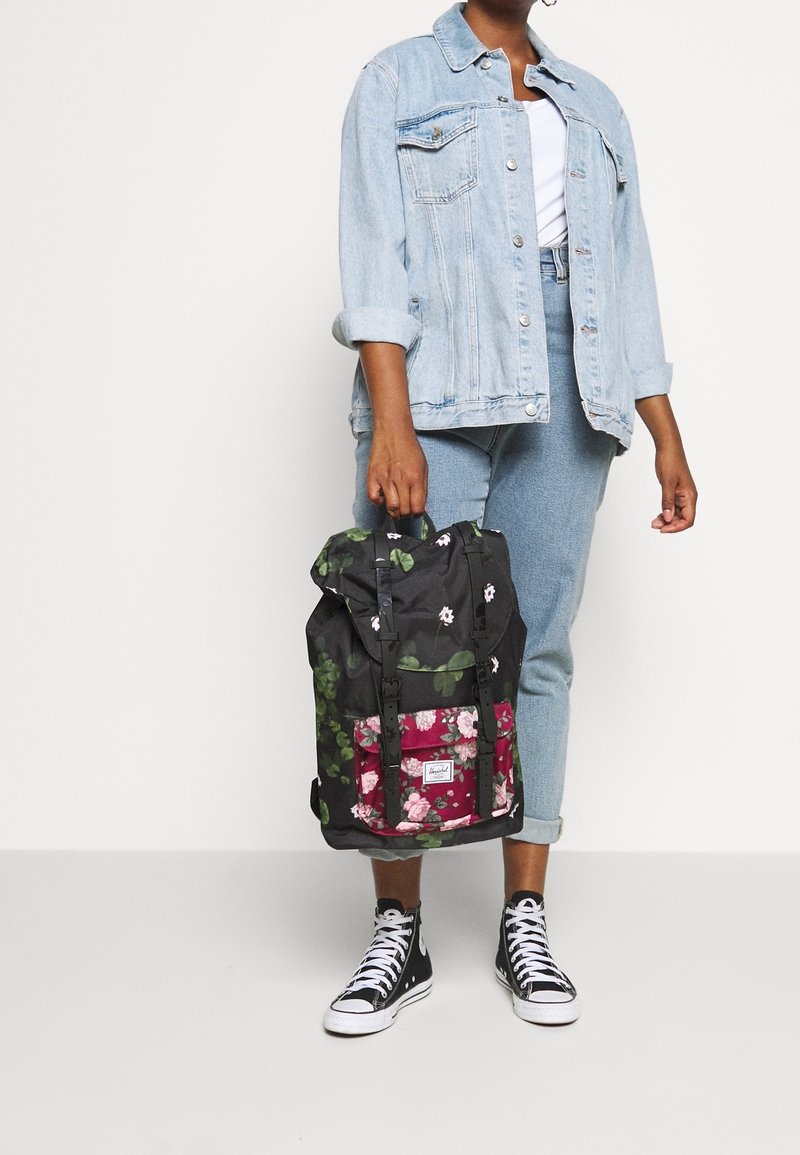Personne portant une veste en jean bleu clair et un jean, tenant un sac à dos noir à motif floral et portant des baskets montantes noires avec des lacets blancs.