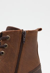 Brown leather boot featuring a textured finish, zip closure, round laces, and a contrasting dark brown section at the base.