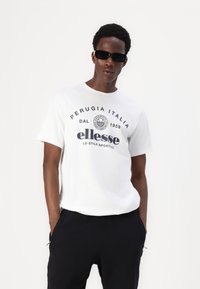 Bílý bavlněný tričko s námořnickým potiskem, na kterém je nápis "Perugia Italia 1959" a logo "ellesse", doplněné černými kalhotami a tmavými slunečními brýlemi.