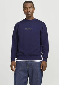 Sudadera marinera con cuello redondo y puños acanalados. Presenta un gráfico blanco de "Originals Studio" en el pecho. La tela es de algodón suave.